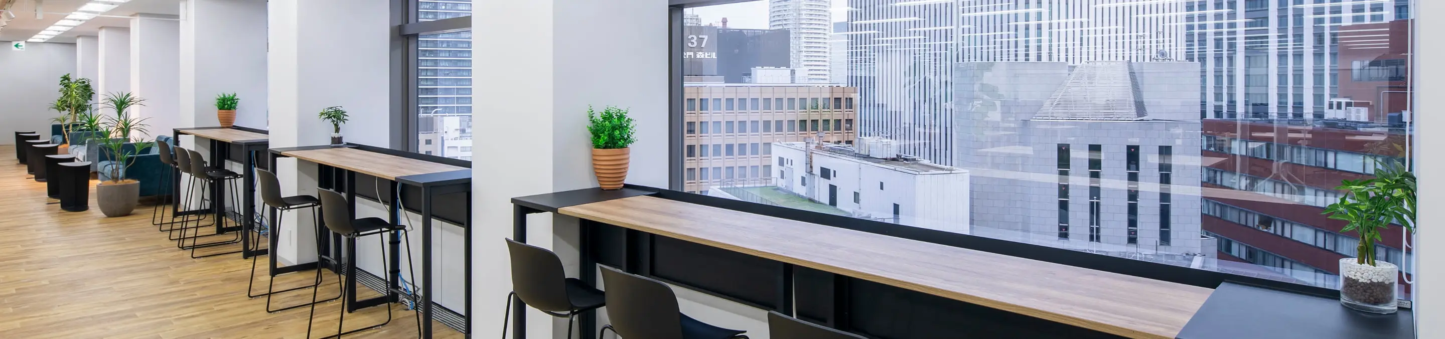 窓際のカウンターとハイスツール、観葉植物が配置された明るいモダンなオフィスのラウンジエリア