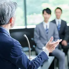 窓から光が差し込む会議室で、スーツ姿の人物が話し、複数の参加者が聞いている様子