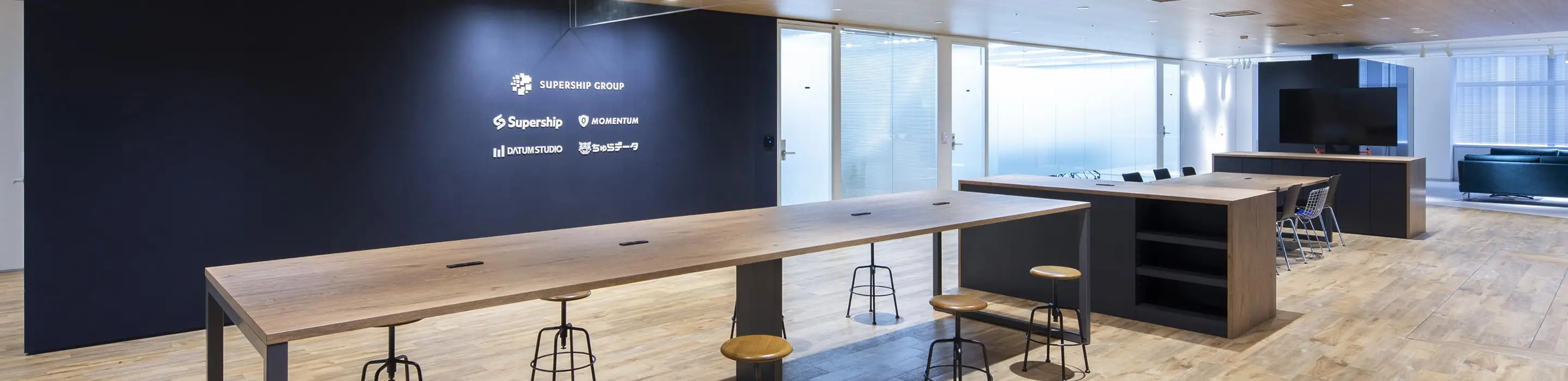 青い壁にロゴが並び、共有テーブルとガラス張りの会議室が配置されたモダンなオフィス空間