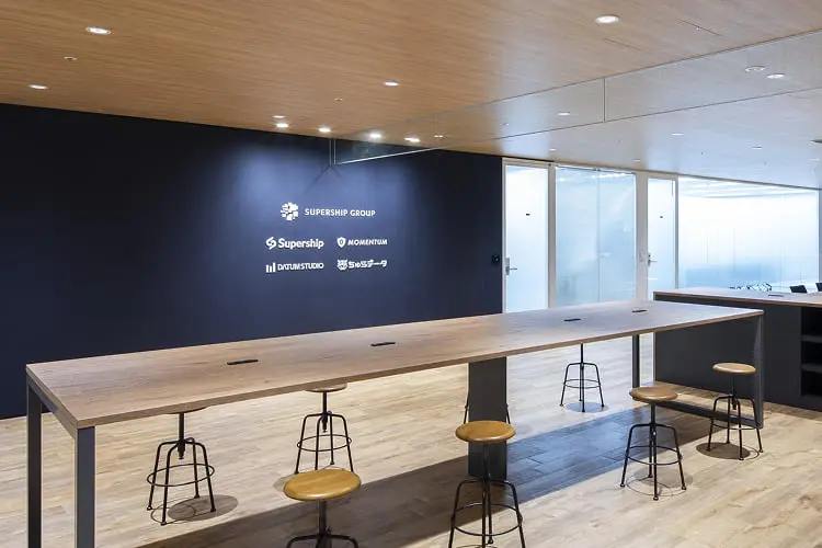 青い壁にロゴが並び、共有テーブルとガラス張りの会議室が配置されたモダンなオフィス空間