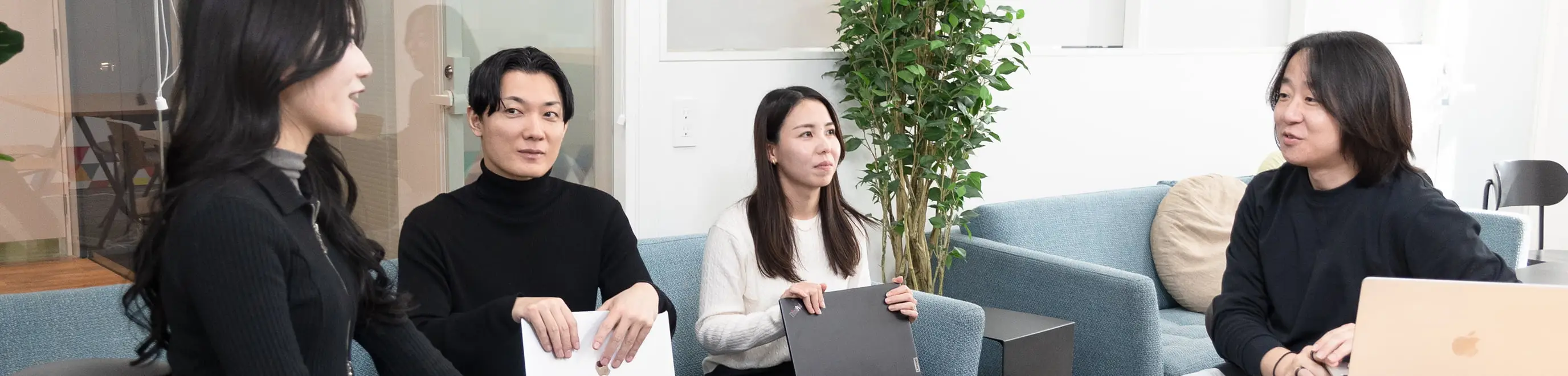 明るいオフィスラウンジで、ソファとテーブルを囲みノートパソコンや資料を見ながら話し合う4人のビジネスパーソン