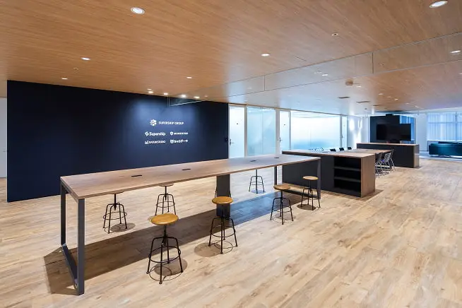 青い壁にロゴが並び、共有テーブルとガラス張りの会議室が配置されたモダンなオフィス空間