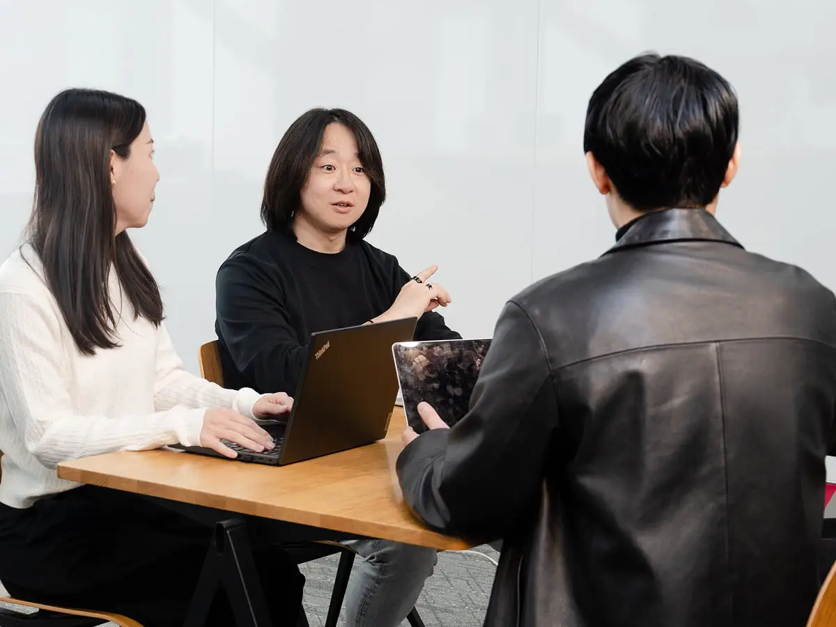 会議テーブルでノートパソコンとタブレットを使いながら話し合う3人のビジネスパーソン