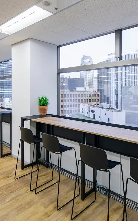 都市の景色を望む窓際の木製カウンターデスクとバースツールが並ぶ明るいオフィススペース