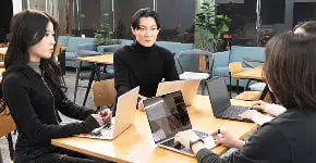 木製テーブルを囲んでノートパソコンで作業する3人のビジネスパーソン